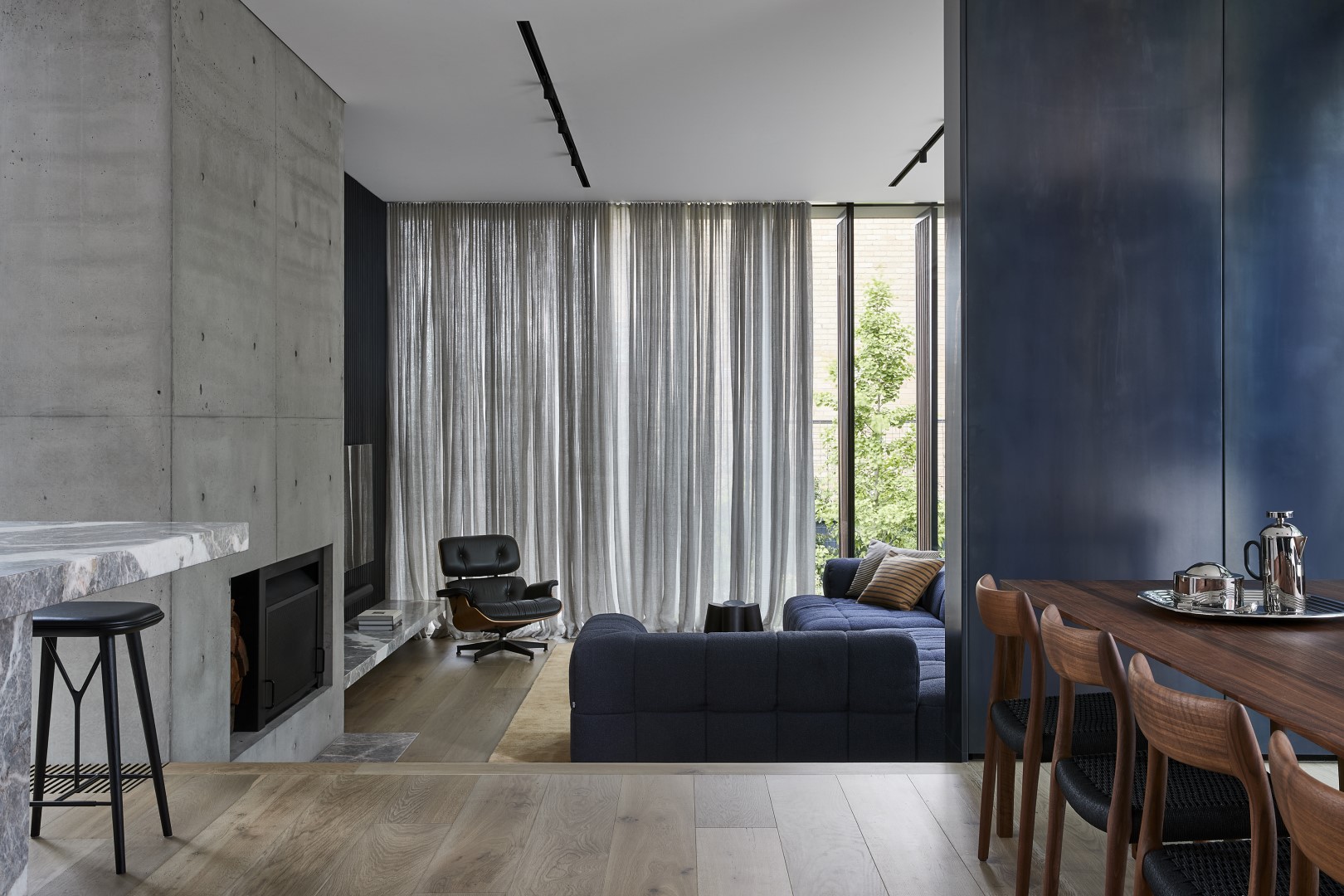 A modern living room featuring warm grey timber flooring with oversized oak floorboards, a concrete fireplace, and a sleek blue sofa. Floor-to-ceiling sheer curtains allow natural light to filter through, enhancing the airy atmosphere.